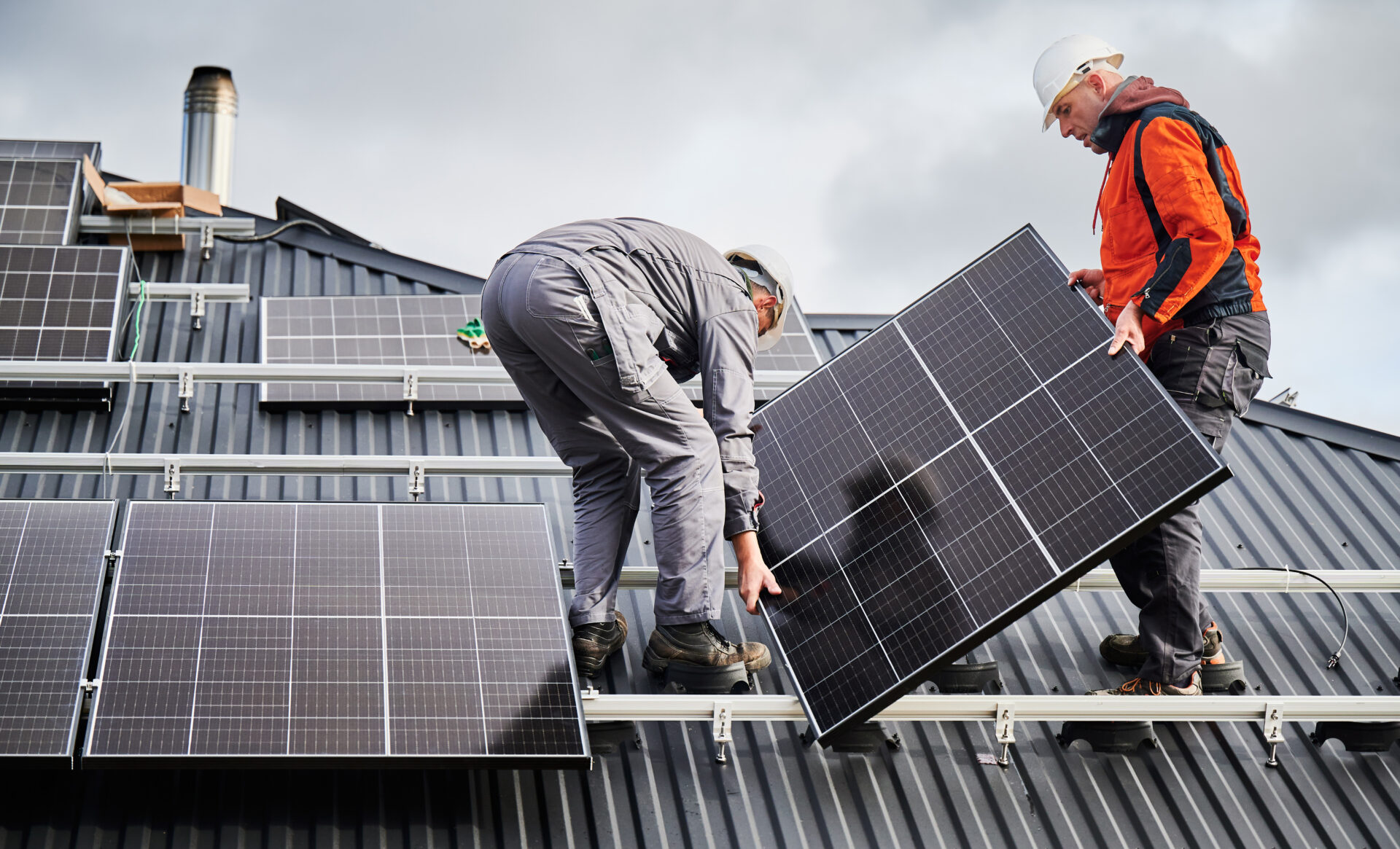 Engineers building solar panel system on roof of house. Men workers in helmets carrying photovoltaic solar module outdoors. Concept of alternative and renewable energy.